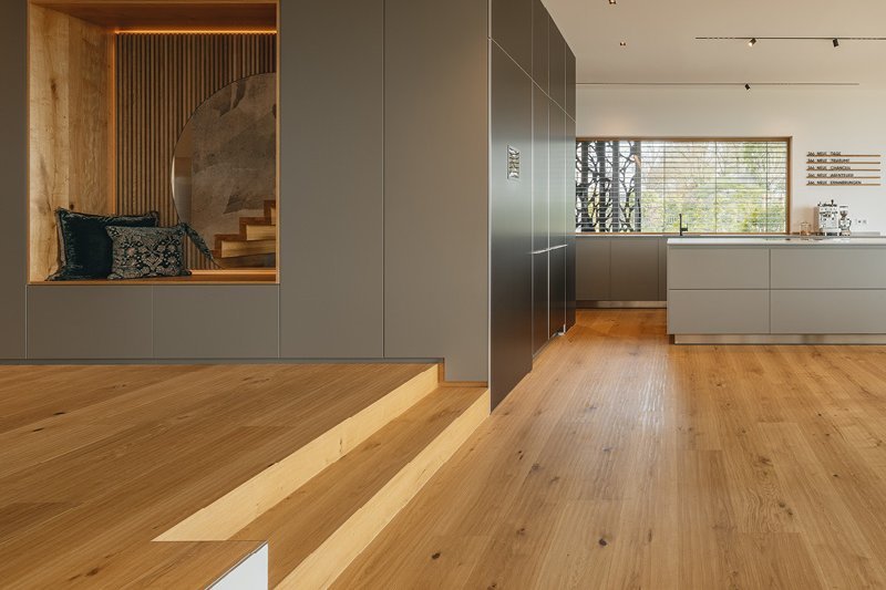 [Translate to EN:] Moderne Küche mit warmem Eichenparkett in Landhausdiele-Optik. Eichenstufen verbinden die Küche mit dem angrenzenden Zimmer. Graue Küchenfronten ergänzen das elegante Design.