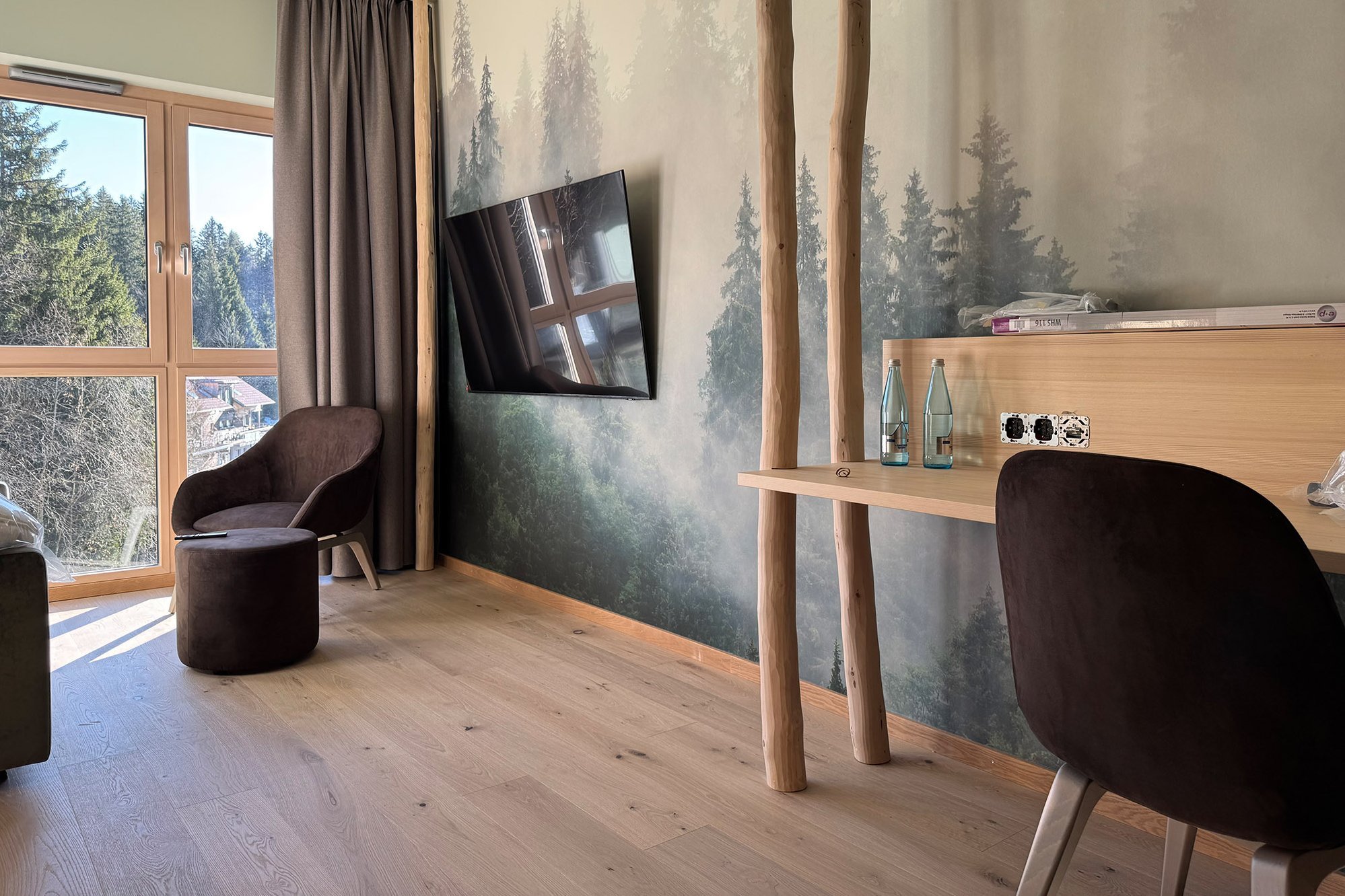 Hotelzimmer mit hellem Eichenparkett, Waldtapete, Holzfensterblick auf Baumwipfel, braune Stoff-Sitzmöbel und Holzmöbel.