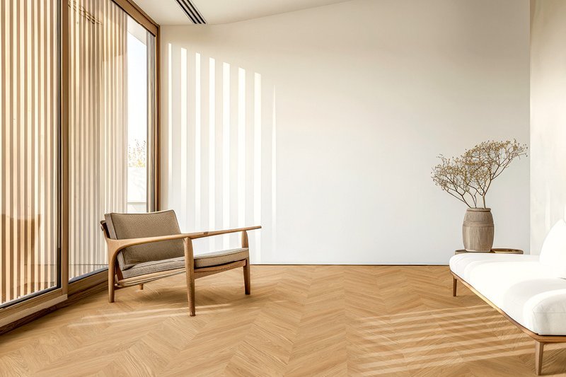 Heller Wohnraum mit hohen weißen Wänden und Eichenparkett im Französischen Fischgrätmuster. Der Raum ist mit einem Holzsessel, einer Vase mit Trockenblumen und einer weißen Couch mit Holzbeinen eingerichtet.