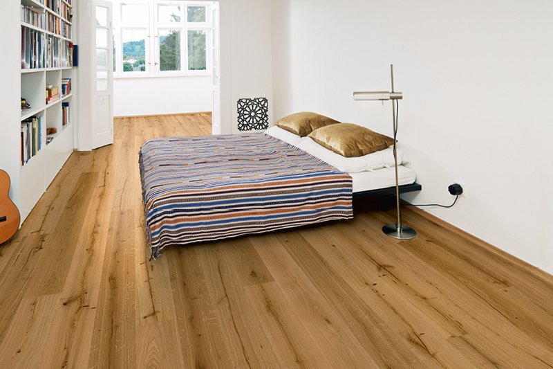 Schlafzimmer mit großer Bücherwand und einer Gitarre. Das Zimmer ist mit einem Parkettboden in Eiche ausgestattet und minimalistisch mit einem Bett und bunter Bettwäsche eingerichtet. Der Raum sieht nach einer Altbauwohnung aus.