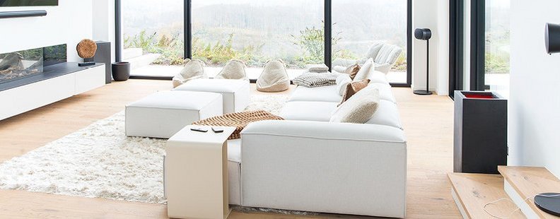 Wohnzimmer in einer Villa mit Parkett aus weißer Eiche, umgeben von einer weißen Sitzlandschaft, einem weißen Fellteppich und schwarzen Akzenten an Möbeln, Fensterrahmen und Kamin.