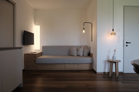 Modern furnished living room with a bed bench with gray linen cover and wood paneling. The floor is covered with rustic, medium brown oak parquet. Black industrial lights provide modern lighting.
