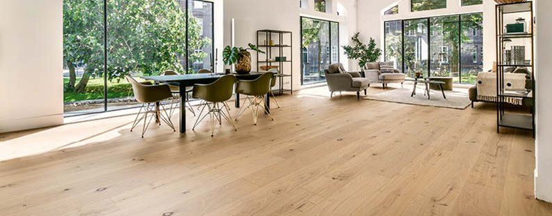 Offener Raum mit bodentiefen Fenstern und Blick auf Bäume. Moderne Möbel, darunter ein Esstisch und Sitzgelegenheiten in Grün und Grau, sowie Eichenparkett in Landhausdielen-Optik.