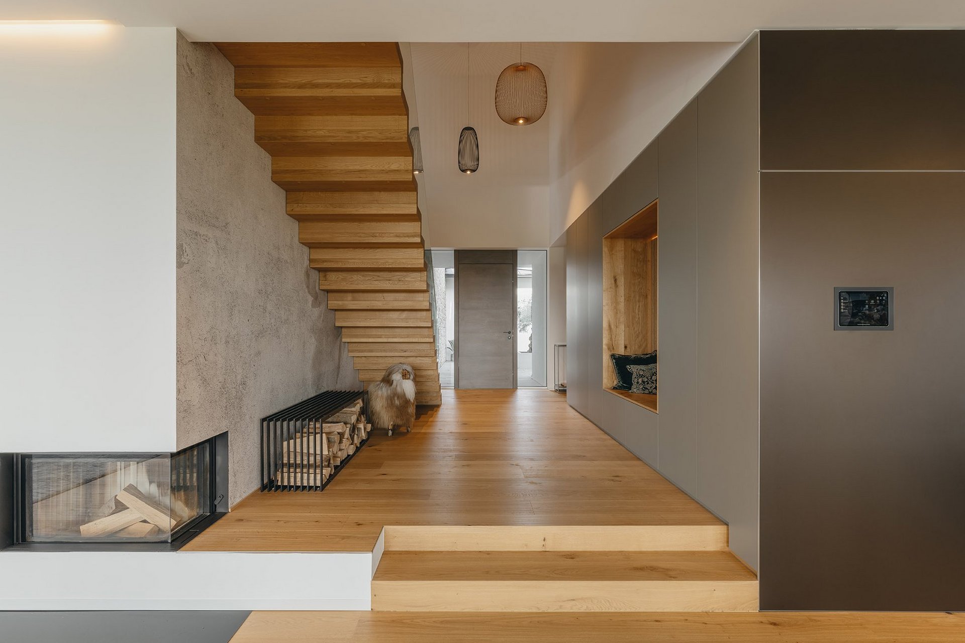 Eingangsbereich eines Hauses mit einer Eichenparkett-Treppe, die in den ersten Stock und in das Wohnzimmer führt. Der Boden ist von einem schönen Kamin und grauen Möbeln umgeben.