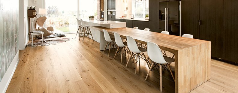 Moderne Küche mit schwarzen Holzfronten. Der Esstisch ist aus Eichenholz und direkt in die Kücheninsel integriert. Am Boden befindet sich ebenfalls Eichenparkett mit rustikaler Optik.