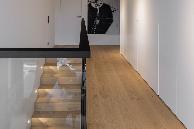 Flur und Treppenaufgang mit Eichenparkett im Landhausdielen Design. Die Holztreppe ist von einem Glasgeländer mit schwarzem Rand umgeben.
