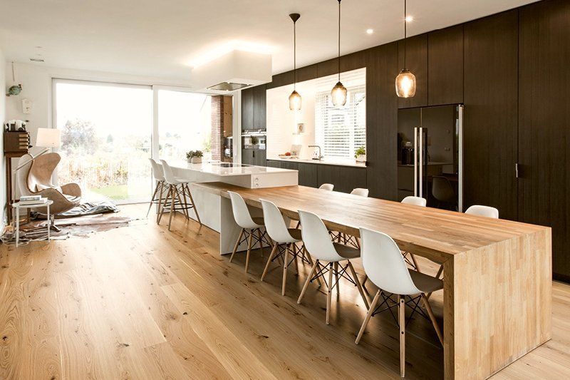 Küche mit schwarzen Holzfronten bis zur Decke. Davor sieht man eine Kücheninsel in weiß mit einem direkt anschließenden Eichenholztisch und weißen Esszimmerstühlen. Am Boden liegt ein rustikales Eichenparkett.