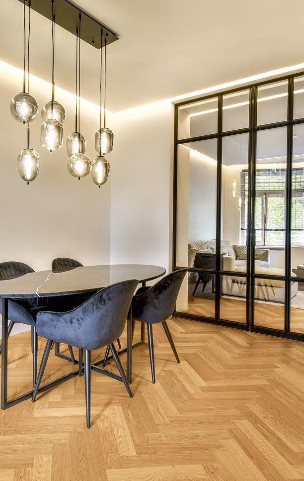 Esszimmer mit Rauchglas Hängeleuchte über einem Marmor-Esstisch und Samtstühlen. Im Raum befindet sich Eichenparkett als Fischgrät verlegt und eine schwarze Industrial-Türe führt in das Wohnzimmer.