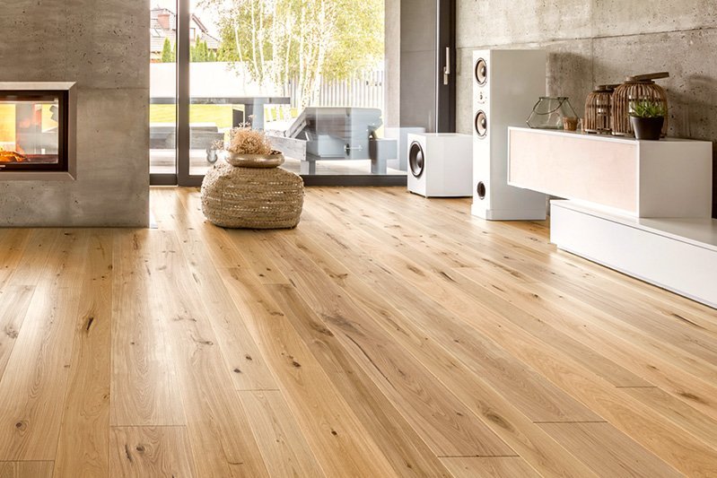 Floor with oak planks surrounded by exposed concrete walls. There is a pouf on the floor and there are white shelves on the walls