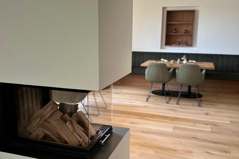 Ansicht eines modernen Kamins auf eine Esstischgruppe mti Holztisch und grünen Stühlen. Im Raum liegen Landhausdielen in Eiche