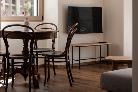 Hotel room with a small, round wooden table and dark brown retro chairs with curved backrests. The floor is covered with oak planks. A television hangs on one wall with an open shelf underneath.