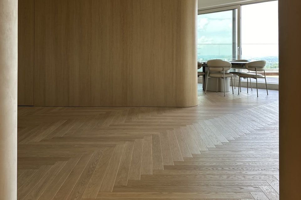 BILAflor 1000 Oak Natur Bianca A bright living room with large white oak herringbone parquet flooring, wood-paneled walls, and seating.