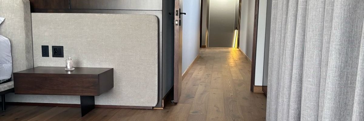 Schlafzimmer mit mittelbraunem Eichenparkett rustikal. Ein beiges Bett, beige Vorhänge und ein grauer Teppich. Eine dunkle Holzwand und chromfarbene Leuchten runden das Raumkonzept ab. Man sieht durch die offene Tür auch in einen Flur, wo auch das Parkett verlegt ist.