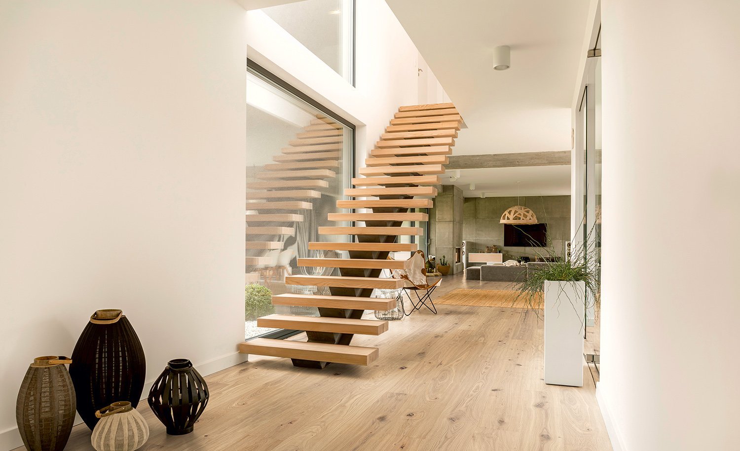 Wohnraum mit freischwebender Stiege aus Eichenholz und rustikalem Eichenparkett am Boden. Der Flur führt in ein Wohnzimmer mit Sichtbetonmauer, grauem Sofa und natürlichen Dekoelementen.