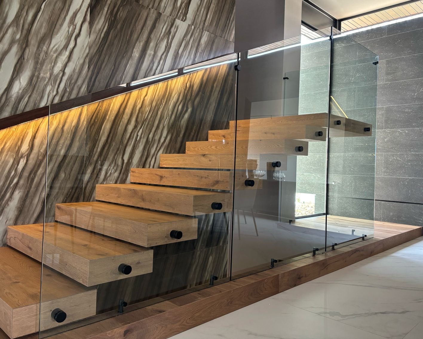 Kragarmtreppe Landhausdiele Eiche Gozo Vorraum mit großen wilden Steinplatten und einer Treppe aus Eichenparkett. das Geländer ist aus Glas und ein schwarzer Handlauf mit integrierter LED-Beleuchtung führt nach oben. Am Vorhausboden liegen weiße Marmorfliesen.