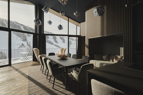 Modernes Hotelzimmer mit großer Fensterfront und Blick auf den Hinterstoder Gletscher, der mit Schnee bedeckt ist. Der Raum ist mit gräulichem Eichenparkett ausgelegt, während schwarze und graue Möbel wie Esstisch und Sessel eine elegante Atmosphäre schaffen. Chromfarbene Pendelleuchten hängen von der Decke und verleihen dem Raum ein modernes Flair.