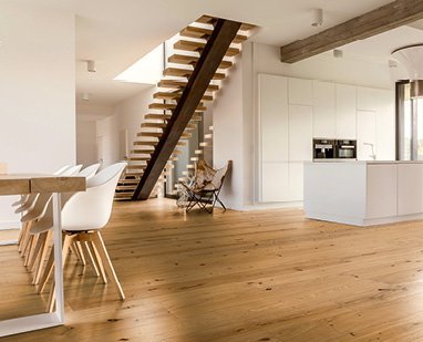 Offene Küche mit weißen Schränken und Kücheninsel. Im Raum befindet sich auch eine Holztreppe und ein Tisch sowie Parkettboden aus Eiche