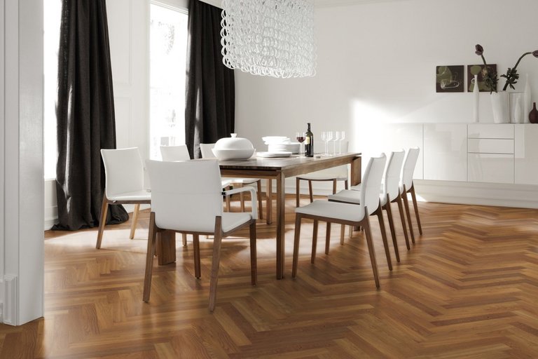 Klassisches Esszimmer mit Fischgrät Parkett in Eiche. Weiße Stühle mit passenden Holzbeinen umgeben einen gedeckten Holztisch. Ein weißes Sideboard mit Dekoartikel ziert die Wand und eine weiße lange Pendelleuchte mit aussergewöhnlichem Design hängt von der Decke. Neben den hohen Fenstern hängen dunkelbraune Vorhänge.