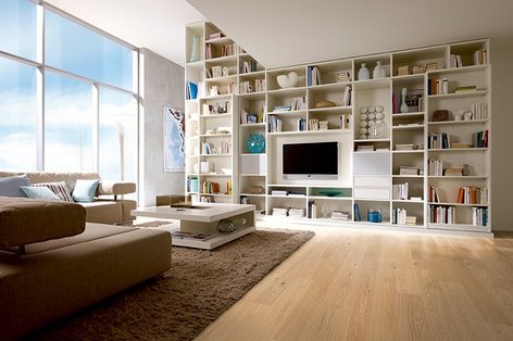 Wohnzimmer mit Bücherwand und hellem Eichenparkett, großflächigen Fenstern mit Ausblick in den Himmel.