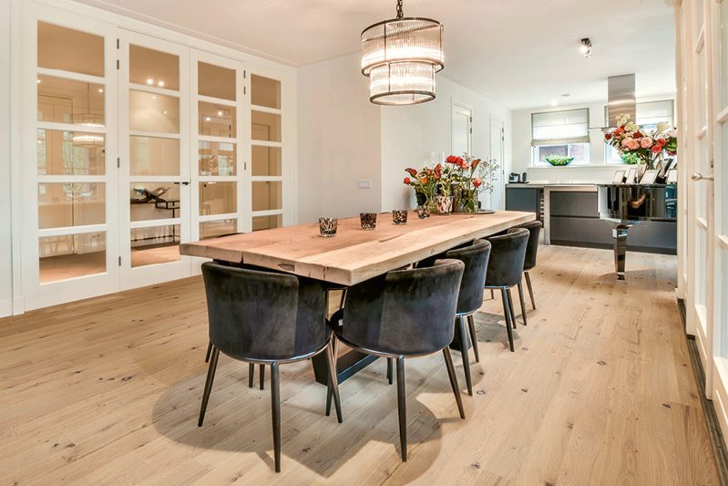 Elegant dining room with gray velvet chairs on grayish oak floorboards. There is also a piano and numerous bouquets of flowers in the room. A noble chandelier hangs above the dining table.