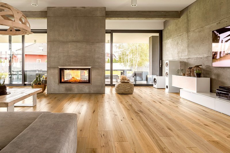 Modernes Wohnzimmer mit Sichtbeton-Kamin und -Mauer. Der Boden ist mit Eichenparkett in rustikaler Optik verlegt. Bodentiefe Fenster bieten einen Ausblick auf den Garten und benachbarte Wohnhäuser.